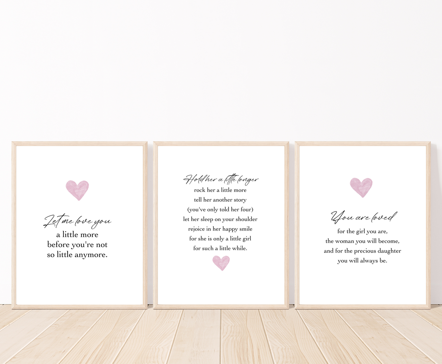 Three frames for a baby girl’s room graphics are placed on a white wall and parquet flooring. The first one shows a pink heart with a piece of writing below that says: Let me love you, a little more before you’re not so little anymore. The middle one has a pink heart below the piece of writing. The last one shows a pink heart with a piece of writing below that says: You are loved for the girl you are, the woman you will become, and for the precious daughter you will always be.