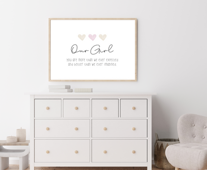 A picture showing a white dresser with a little girl’s graphic hanging on the wall right above the latter. The graphic is showing three pink and beige hearts with a piece of writing below that says: Our girl you are more than we ever expected, and better than we ever imagined.