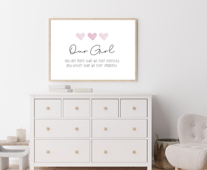 A picture showing a white dresser with a little girl’s graphic hanging on the wall right above the latter. The graphic is showing three pink hearts with a piece of writing below that says: Our girl you are more than we ever expected, and better than we ever imagined.
