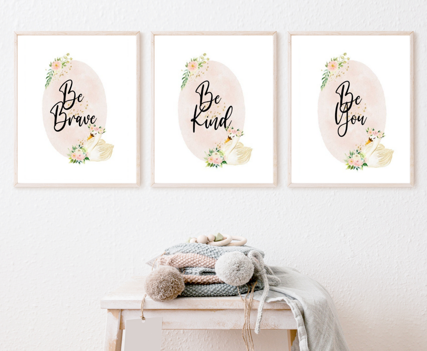 An image showing three graphics hanging on the wall right above a piece of furniture. The three are identical showing a baby pink oval shape decorated with flowers on the top and the bottom of the shape and beside the flowers at the bottom there is a cute goose illustration. The first graphic says” Be Brave”, the second “Be Kind”, and the third “Be You”.