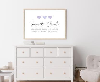 A picture showing a white dresser with a little girl’s graphic hanging on the wall right above the latter. The graphic is showing three purple blue hearts with a piece of writing below that says: Sweet girl you are more than we ever expected, and better than we ever imagined.