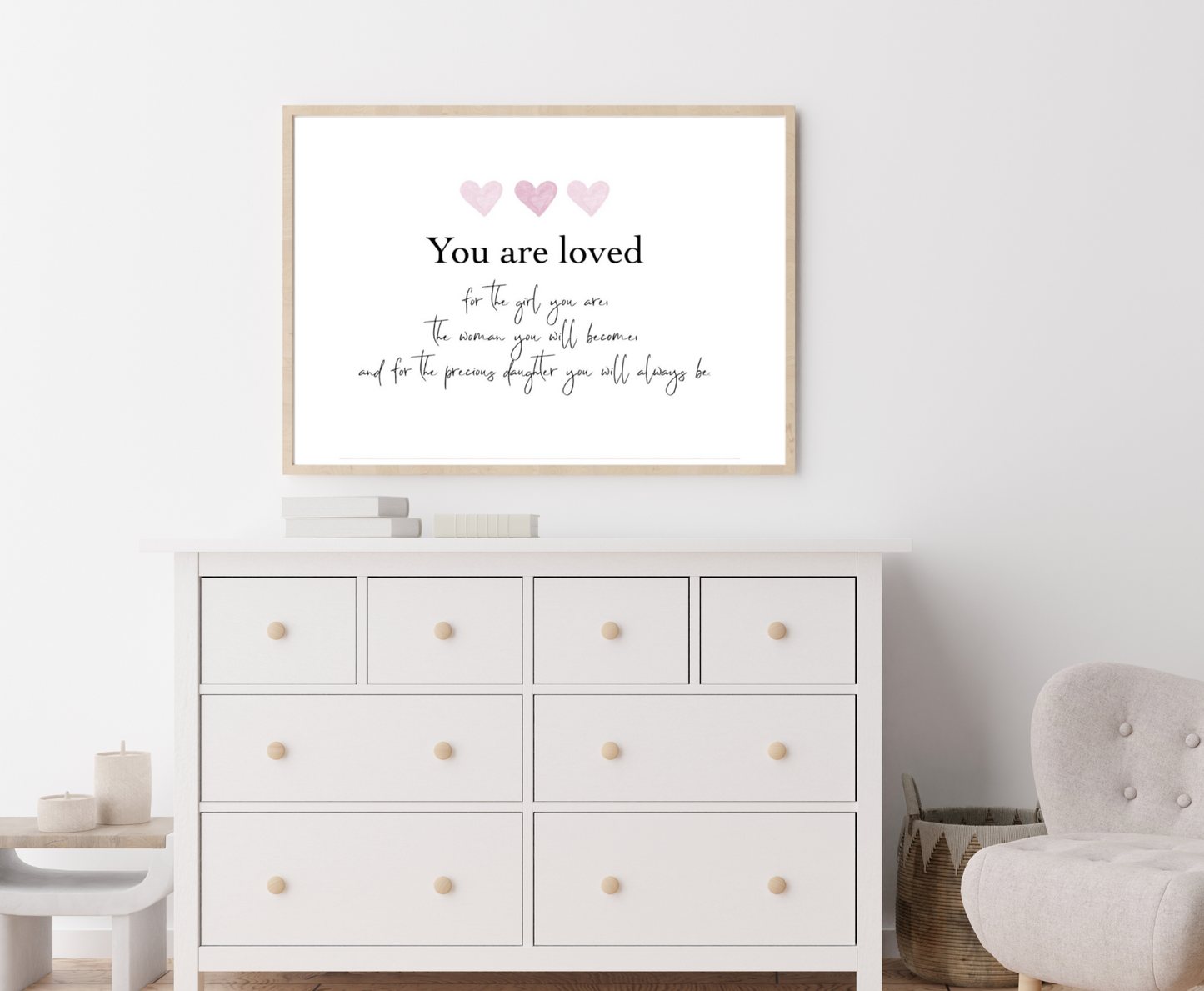 A picture showing a white dresser with a little girl’s graphic hanging on the wall right above the latter. The graphic is showing three pink hearts with a piece of writing below that says: You are loved for the girl you are, the woman you will become, and the precious daughter you will always be.