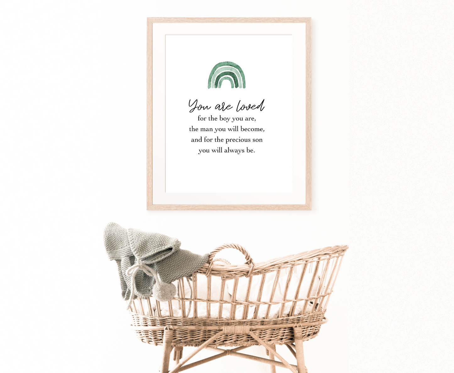 An image showing a cradle with a frame hung on the wall behind it. The frame shows a graphic for a little boy’s room that has an ombre green rainbow design and includes the following “You are loved, for the boy you are, the man you will become, and for the precious son you will always be.” 