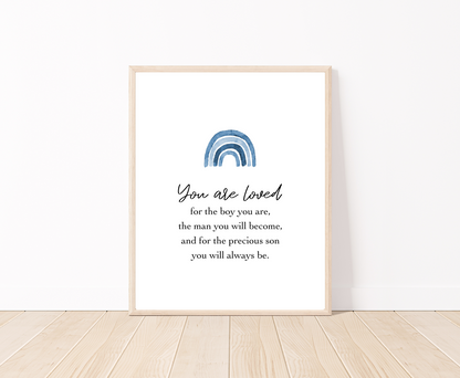 A digital poster that is placed on a white wall and parquet flooring and has a blue rainbow at the top with a piece of writing that says: “You are loved for the boy you are, the man you will become, and for the precious son you will always be.”