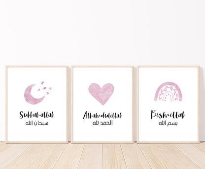 Three frames showing graphics for a baby girl’s room that are placed on a white wall and parquet flooring. The first one shows a small pink moon and three small stars with a piece of writing below that says: “Subhanallah”. The middle graphic shows a pink heart with a piece of writing below that says: “Alhamdulillah”. The last one shows a pink rainbow with a piece of writing below that says: “Bismillah”.