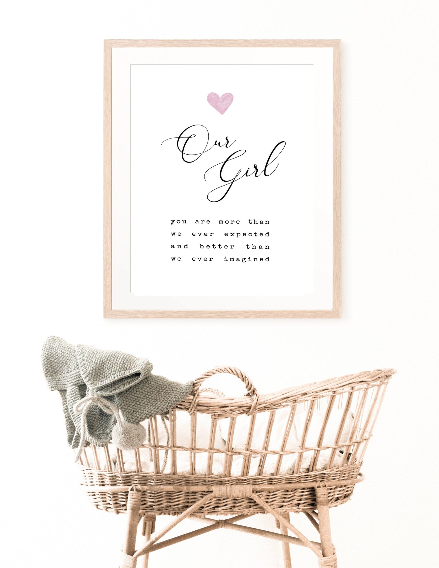 A digital print is hung above a baby’s cradle. The digital print has a pink heart at the top, and a piece of writing that says: “Our girl, you are more than we ever expected and better than we ever imagined.”