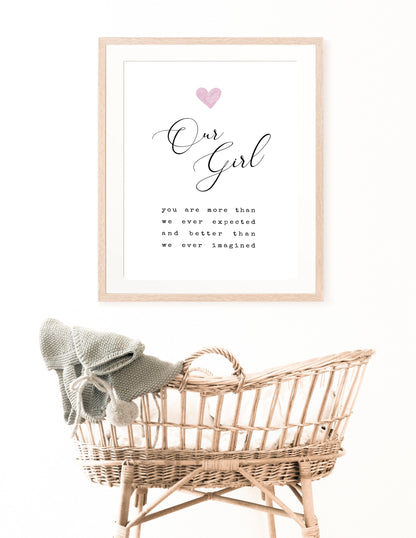 A digital print is hung above a baby’s cradle. The digital print has a pink heart at the top, and a piece of writing that says: “Our girl, you are more than we ever expected and better than we ever imagined.”