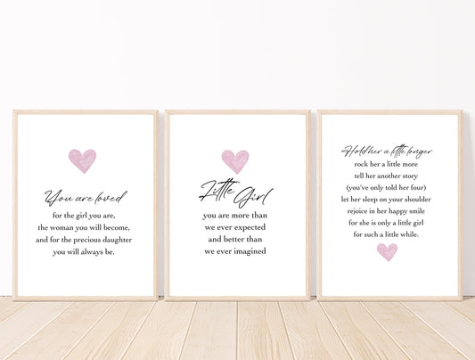 Three frames for a baby girl’s room graphics that are placed on a white wall and parquet flooring. The first one shows a pink heart with a piece of writing below that says: You are loved for the girl you are, the woman you will become, and for the precious daughter you will always be. The middle graphic shows a pink heart with a piece of writing below that says: little girl you are more than we ever expected and better than we ever imagined. The last one has the heart below the piece of writing.