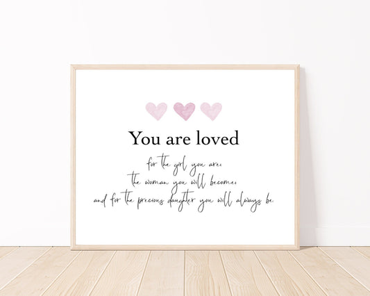 A digital poster is placed on a white wall and parquet flooring and has three pink hearts at the top with a piece of writing that says: “You are loved for the girl you are, the woman you will become, and for the precious daughter you will always be.”