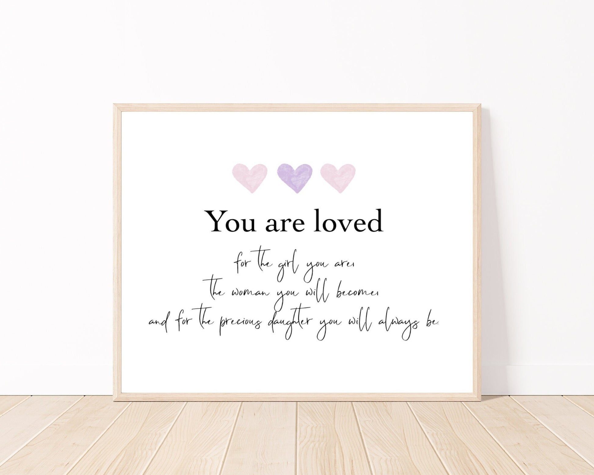 A little girl’s room digital poster that is placed on a white wall and parquet flooring and has three hearts at the top with a piece of writing that says: “You are loved for the girl you are, the woman you will become, and for the precious daughter you will always be.”