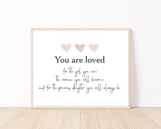 A little girl’s room digital poster that is placed on a white wall and parquet flooring and has three hearts at the top with a piece of writing that says: “You are loved for the girl you are, the woman you will become, and for the precious daughter you will always be.”