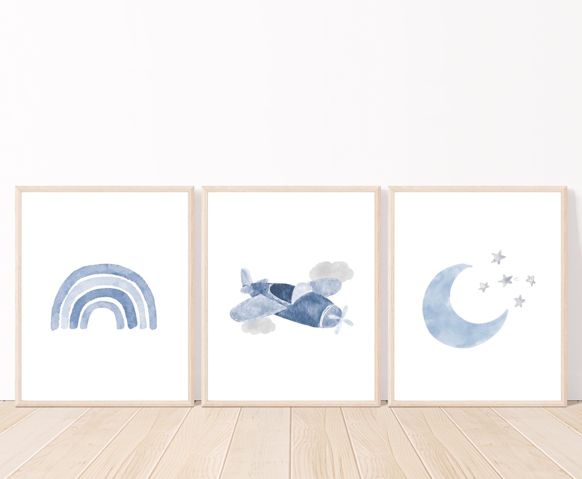 An image showing three digital prints placed on a white wall and parquet flooring. The first one shows a baby blue rainbow design. The second one shows a blue airplane and two gray clouds. The third one shows a baby blue crescent and some tiny gray stars beside it.