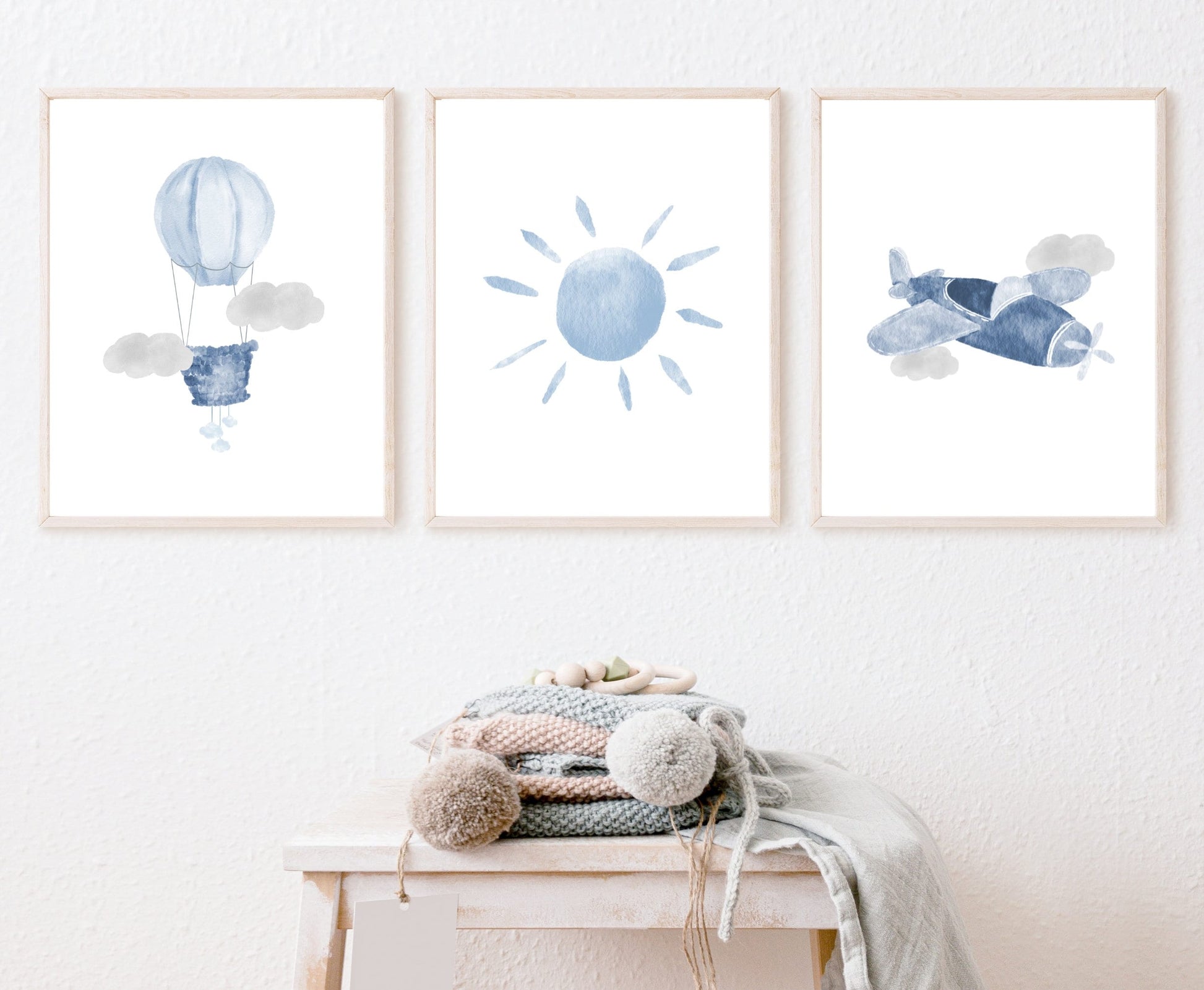 An image showing three digital prints that are hung on a wall. The first one shows a baby blue air balloon with two grey clouds design. The second one shows a baby blue sun. The third one shows a baby blue airplane and two gray clouds.